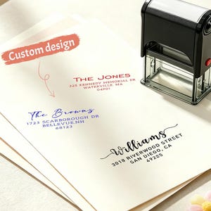 Op de afbeelding: Een zwart-witte adresstempel ligt op een stapel enveloppen met gedrukte adressen. De enveloppen tonen de namen en adressen van "The Jones", "The Browns" en "Williams". Een label met een aangepast ontwerp is zichtbaar.