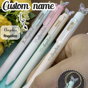Puede incluir: Bolígrafos de colores pastel con el nombre "Angelica" grabado. Cada bolígrafo está coronado con una mariposa decorativa. La imagen también incluye el texto "Custom name" y "A butterfly that spins."