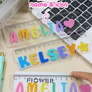 May include: Transparent acrylic rulers featuring the names "AMELIA" and "KELSEY" in bright, multi-coloured letters. A ruler with the name "AMELIA" and the word "FLOWER" is also shown. The rulers have centimetre markings and are decorated with icons.