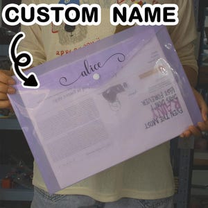 Peut inclure: Une pochette de documents en plastique transparent avec un rabat lavande, portant le nom "Alice" en cursive. La pochette est fermée par un bouton-pression blanc et contient des papiers. D'autres pochettes en turquoise, corail et bleu sont présentées en bas.