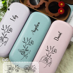 May include: Three pastel-coloured personalised glasses cases: lavender, teal, and pink. Each case has a name in cursive script and a floral design with a letter. The top of the image shows a brown glasses case with sunglasses.