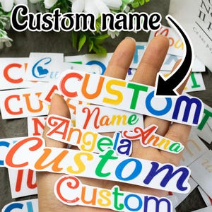 Può includere: Una mano mostra diversi adesivi con nomi personalizzati con un design a gradiente arcobaleno. Gli adesivi presentano la parola "Custom" e vari nomi in diversi caratteri. Lo sfondo include fiori bianchi e adesivi aggiuntivi.