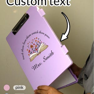 Op de afbeelding: Een lichtpaarse clipboard met een metalen clip, met de tekst "Custom text" bovenaan. De clipboard heeft een bloemenontwerp met de woorden "reach them love them watch them grow" en "Mrs. Smith".