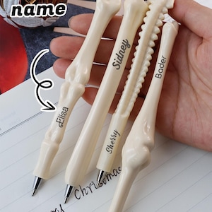 Puede incluir: Cuatro bolígrafos con forma de hueso en blanco roto, cada uno con punta plateada y nombre personalizado. Los bolígrafos se sostienen sobre un cuaderno con la frase "Merry Christmas" escrita. La imagen también incluye el texto "Custom name".