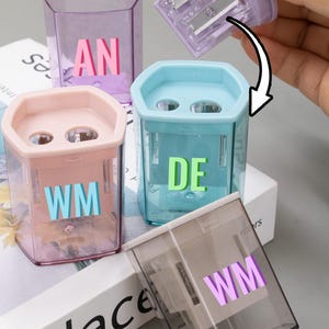 May include: A selection of transparent pencil sharpeners in various colours, including pink, blue, purple, and grey. Each sharpener has a different letter combination. One sharpener's top is being removed, showing the blades.