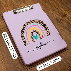 May include: A lavender clipboard with a rainbow design and the name "Sophia". The rainbow features a leopard print section, with pink, yellow, and blue arcs. The clipboard measures 12.79 inches by 9.25 inches.