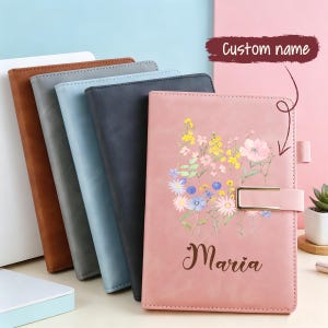 May include: A collection of four rectangular notebooks with smooth covers in brown, grey, light blue, and dark grey, plus a pink notebook featuring a floral print, the name Maria in script, and a gold metal clasp closure on the front cover.