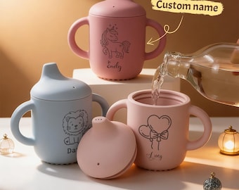 May include: A collection of personalized baby sippy cups in pink and blue. The pink cups feature unicorn and heart designs, while the blue cup has a lion illustration. Each cup has a handle and a custom name. The text "Custom name" is also visible.