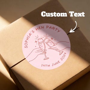 May include: A round pink sticker with the text "Sophia's Hen Party" and "14th June 2024" in red. The sticker features two champagne glasses clinking, with hands holding them. The sticker is on a brown package.