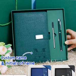 May include: A teal journal and pen set presented in a gift box. The journal features a strap closure with a silver detail. The text "Custom name & initials of name" is visible. Additional sets in blue and black are shown. A gift bag is in the background.