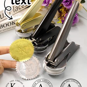 Op de afbeelding: Drie metalen bibliotheekzegels in goud, zwart en zilver, met aangepaste tekst. De afbeelding bevat goud- en zilverkleurige reliëfzegels met de tekst "FROM THE LIBRARY OF" en initialen. De tekst "Custom text" is ook zichtbaar.