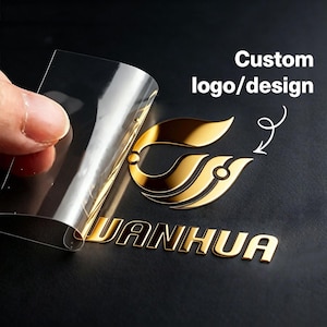 May include: A gold metallic custom logo sticker with the text WANHUA, featuring a stylised leaf-like emblem. A clear transfer film is being peeled back from the surface, revealing the reflective gold finish of the adhesive decal design.
