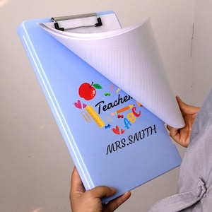 May include: A light blue clipboard with a silver clip holding white paper. The clipboard is decorated with a red apple, hearts, and the word "Teacher" with the name "MRS. SMITH".