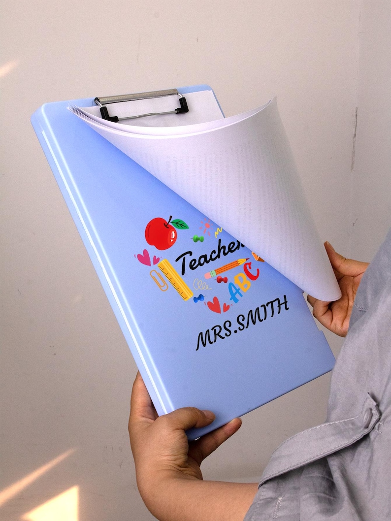 May include: A light blue clipboard with a silver clip holding white paper. The clipboard is decorated with a red apple, hearts, and the word "Teacher" with the name "MRS. SMITH".