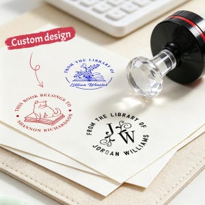 May include: A custom design stamp with a clear crystal handle and black ink pad. Three stamped designs are visible: "From the Library of Lillian Winstet", "This Book Belongs to Shannon Richardson", and "From the Library of Jordan Williams".