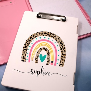 May include: A white clipboard with a rainbow and leopard print design, the name "sophia" is written in cursive. The clipboard is open, revealing a pink interior and a stack of white paper. A silver clip holds the paper in place.