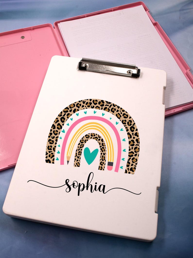 May include: A white clipboard with a rainbow and leopard print design, the name "sophia" is written in cursive. The clipboard is open, revealing a pink interior and a stack of white paper. A silver clip holds the paper in place.