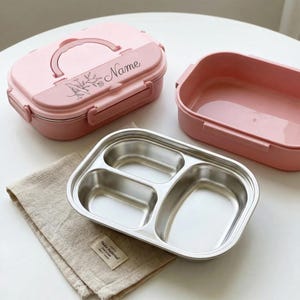 May include: A pink lunch box with a handle and the word "Name" printed on the lid. The lunch box is open, revealing a stainless steel divided tray and a pink container. A beige cloth is in the foreground.