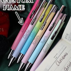 Puede incluir: Un conjunto de bolígrafos personalizados en colores pastel, incluyendo rosa, azul y amarillo, con detalles plateados. Cada bolígrafo está grabado con un nombre diferente. También se ve un bloc de notas con la frase "Merry Christmas".