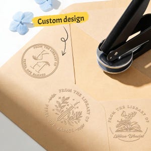 May include: A custom design embosser with three different stamp designs on brown envelopes. The stamps feature text and illustrations, including a sword, an open book, and initials. A black embosser tool is in the image.