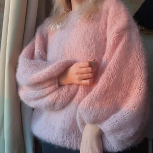 Può includere: Un maglione rosa chiaro oversize con maniche lunghe e voluminose. Il maglione è realizzato in un materiale morbido e soffice e ha uno scollo tondo. Le maniche si raccolgono ai polsi. Il maglione è in tinta unita e sembra essere lavorato a mano.