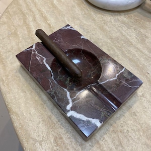 Può includere: Un posacenere rettangolare in marmo bordeaux con venature bianche. Un sigaro marrone è appoggiato nella vaschetta del posacenere. Il posacenere ha una finitura liscia e lucida e una piccola scanalatura per appoggiare un sigaro.