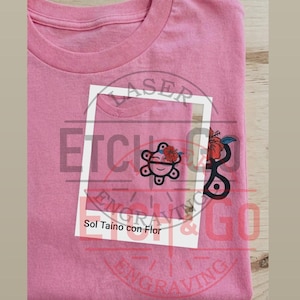 May include: Pink t-shirt with a pocket featuring a graphic design of a turtle with a flower and the text "Sol Taíno con Flor". The shirt is made of a soft material.