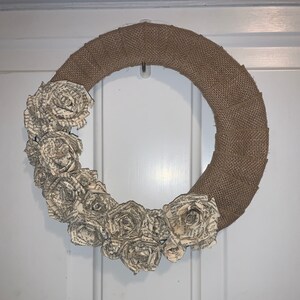 Pode incluir: Uma coroa de juta decorada com rosas de papel feitas de páginas de livros. As rosas estão dispostas em um cacho ao redor da coroa.