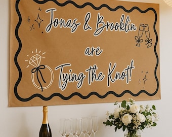 May include: A brown banner with black wavy border and text that reads "Jonas & Brooklin are Tying the Knot!" with illustrations of a diamond ring and champagne glasses. A champagne bottle and six champagne flutes are on a wooden table.