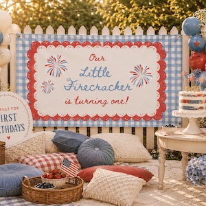 May include: A rectangular banner with a blue and white gingham border and a red scalloped edge, featuring the text Our Little Firecracker is turning one! in blue and red script, flanked by firework illustrations, set against a white fence.