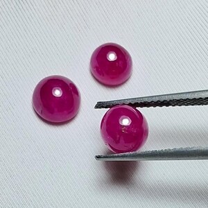 May include: Three round, polished, ruby gemstones. One gemstone is held by tweezers. The rubies are a deep, vibrant red color and have a smooth, reflective surface. The gemstones are on a white background.