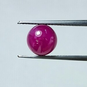 May include: A vibrant, round, polished ruby gemstone held by a pair of silver tweezers. The gemstone is a deep, saturated pinkish-red color, reflecting light with a bright spot on its surface. The background is a plain, light gray.