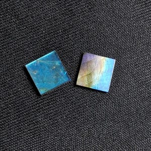 Puede incluir: Dos piedras cuadradas de labradorita con tonos iridiscentes azules, verdes y morados. Las piedras se muestran sobre una superficie oscura y texturizada, mostrando sus colores y patrones naturales. Estas piedras son ideales para la fabricación de joyas.
