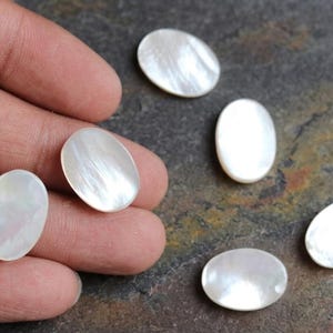 Puede incluir: Cinco cabujones de nácar blanco de forma ovalada y lisa con un acabado lustroso e iridiscente. Dos de los cabujones se sostienen entre los dedos, mientras que otros tres están dispuestos cerca, mostrando sus superficies pulidas y reflectantes.