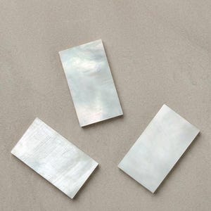 Può includere: Tre pezzi rettangolari di madreperla iridescente con una superficie liscia e lucida. Ogni pezzo mostra una naturale lucentezza bianco perlaceo con sottili variazioni di consistenza e riflessi di luce sulle superfici piatte e sottili.