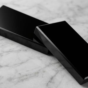 May include: Two rectangular blocks with a smooth, glossy black finish. The blocks have sharp, defined edges and a reflective surface. One block is positioned horizontally in the background, while the other is angled in the foreground.