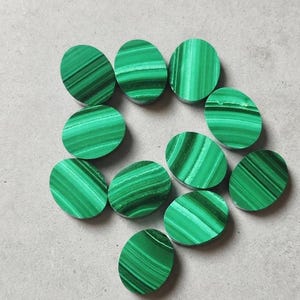 May include: Ten oval-shaped malachite cabochons arranged in a loose cluster. Each piece displays a vibrant green colour with distinct, natural banded patterns of light and dark green stripes across the smooth, polished surfaces of the stones.