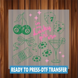 May include: A ready-to-press DTF transfer with a green and pink design. The design includes cherries, dice, poker chips, a martini glass, and the text "in my lucky girl era."