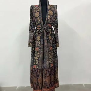 May include: A long, patterned duster coat with a shawl collar and tie waist. The coat features a dark background with floral and geometric designs in shades of gold, red, and brown. The sleeves are long, and the coat reaches the ankles.