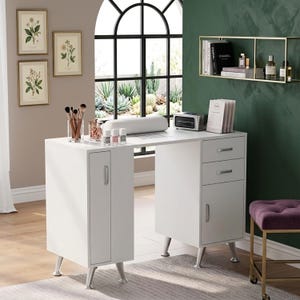 Puede incluir: Una mesa de manicura blanca con reposabrazos incorporado, cajones y un armario. La mesa tiene patas plateadas y está colocada contra una pared verde con estampados botánicos enmarcados. Se ven botellas de esmalte de uñas y brochas de maquillaje.