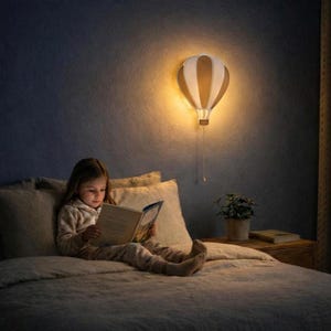 Puede incluir: Una lámpara de pared con forma de globo aerostático, con paneles beige y blancos, ilumina a un niño leyendo en la cama. La lámpara está montada en una pared azul oscuro, proyectando un brillo cálido. El niño lleva un pijama y lee un libro.