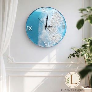 Peut inclure: Horloge murale ronde avec un motif océan bleu et blanc. Le cadran de l'horloge présente des chiffres romains et un motif texturé en forme de vague. L'horloge est fixée sur un mur blanc.