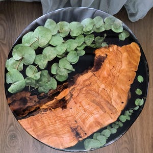 Könnte beinhalten: Runder Tisch mit dunkler Harzoberfläche. Der Tisch hat einen Abschnitt mit hellbrauner Holzmaserung und grüne Eukalyptusblätter, die in das Harz eingebettet sind. Der Tisch steht auf einem Holzboden.