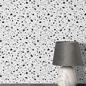 Op de afbeelding: Een grijze tafellamp met een marmerachtige voet en een grijze lampenkap. De achtergrond is een witte muur met een terrazzopatroon van zwarte, grijze en witte chips.