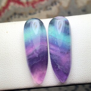 May include: Two polished, teardrop-shaped fluorite gemstones featuring a smooth, glossy finish. The stones display a vibrant gradient of colors, transitioning from deep purple at the pointed base to bright teal and light blue at the rounded top.