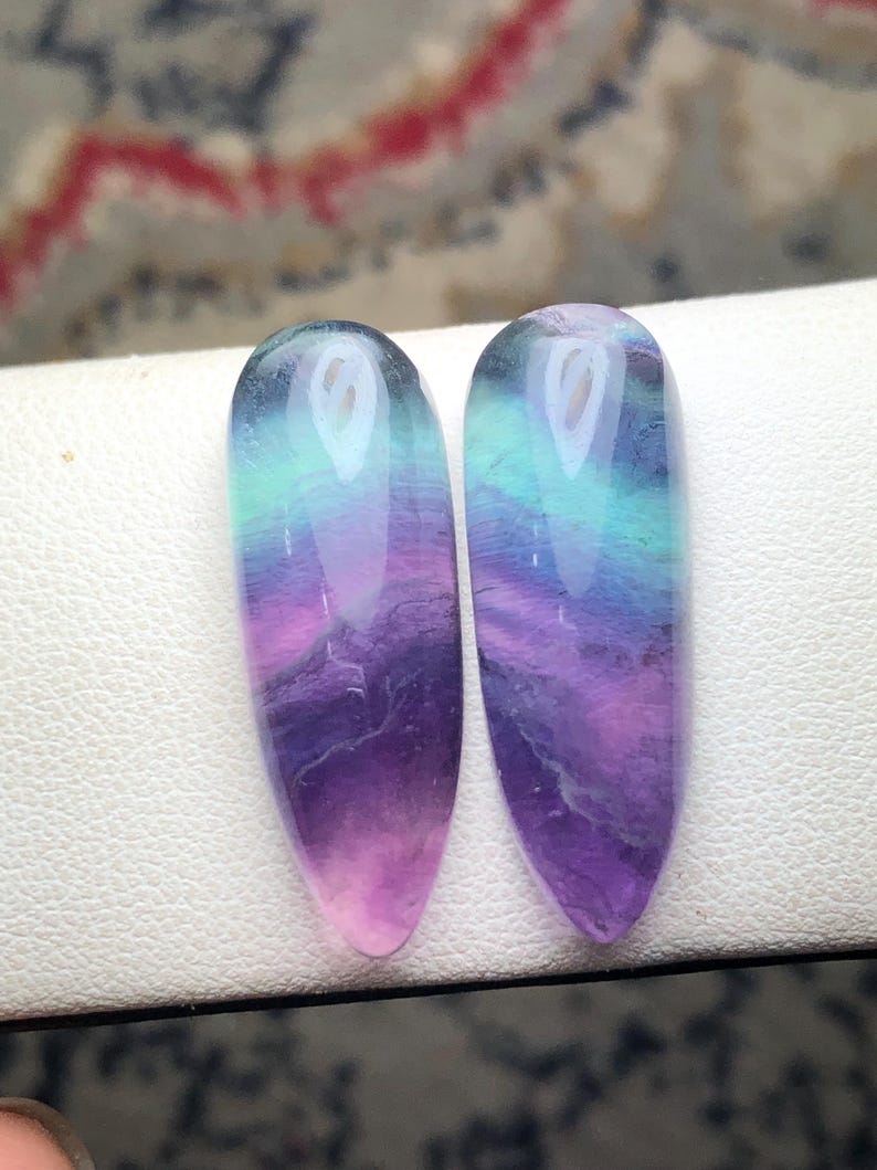 May include: Two polished, teardrop-shaped fluorite gemstones featuring a smooth, glossy finish. The stones display a vibrant gradient of colors, transitioning from deep purple at the pointed base to bright teal and light blue at the rounded top.