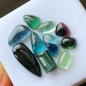 K&ouml;nnte beinhalten: Eine Sammlung polierter Edelsteine in verschiedenen Formen und Farben, darunter Gr&uuml;n, Blau und Schwarz. Die Edelsteine werden auf einer wei&szlig;en Oberfl&auml;che pr&auml;sentiert, die ihre durchscheinenden und reflektierenden Eigenschaften hervorhebt. Die Sammlung umfasst Turmalin und andere Edelsteine.