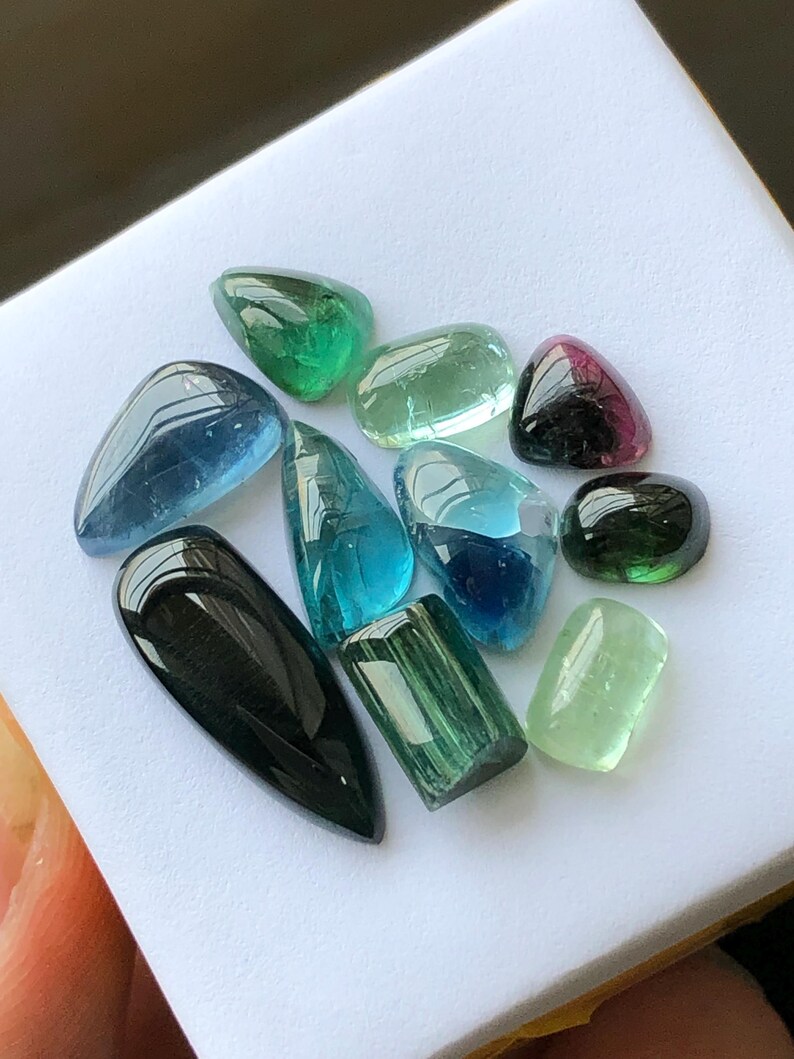 K&ouml;nnte beinhalten: Eine Sammlung polierter Edelsteine in verschiedenen Formen und Farben, darunter Gr&uuml;n, Blau und Schwarz. Die Edelsteine werden auf einer wei&szlig;en Oberfl&auml;che pr&auml;sentiert, die ihre durchscheinenden und reflektierenden Eigenschaften hervorhebt. Die Sammlung umfasst Turmalin und andere Edelsteine.