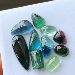 K&ouml;nnte beinhalten: Eine Sammlung polierter Edelsteine in verschiedenen Formen und Farben, darunter Blau, Gr&uuml;n und Lila. Die Edelsteine werden auf einer wei&szlig;en Oberfl&auml;che pr&auml;sentiert, mit einer Vielzahl von Schliffen und Gr&ouml;&szlig;en. Die Edelsteine haben ein reflektierendes, gl&auml;nzendes Aussehen.