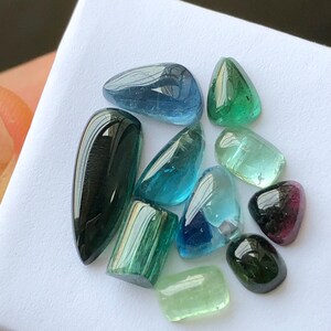 K&ouml;nnte beinhalten: Eine Sammlung polierter Edelsteine in verschiedenen Formen und Farben, darunter Blau, Gr&uuml;n und Schwarz. Die Edelsteine werden auf einer wei&szlig;en Oberfl&auml;che pr&auml;sentiert, die ihre durchscheinenden und reflektierenden Eigenschaften hervorhebt. Die Auswahl umfasst ovale, tropfenf&ouml;rmige und rechteckige Schnitte.